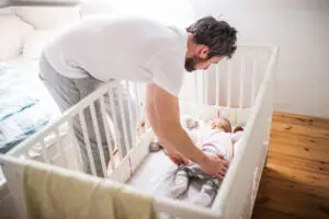 Father putting a sleeping toddler girl into cot at home. - baby sleep consultant
